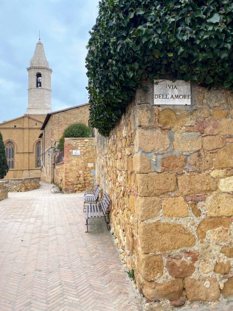 Sign for Via dell'Amore on the stone wall in Pienza, Italy. Ivy above it and a church behind and to the left.
