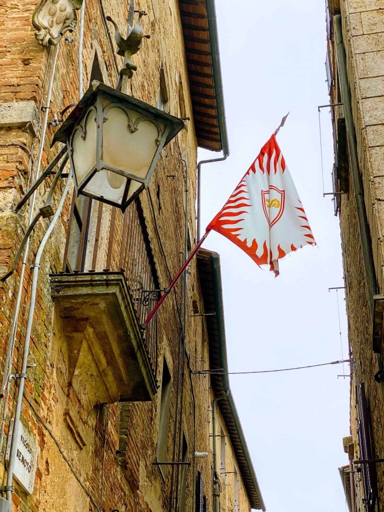 Red and white flag hung from a wall in on a tiny street in the San Donato neighborhood in Montepulciano Italy. The buildings are red brick and stone and there is also an old iron lamp and a stone crest of the Medici family.