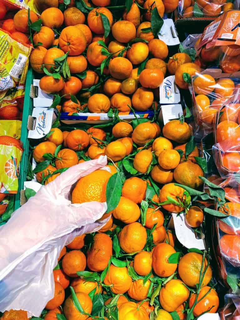 A hand with a plastic glove chooses a clementine from a large pile at an Italian grocery store.