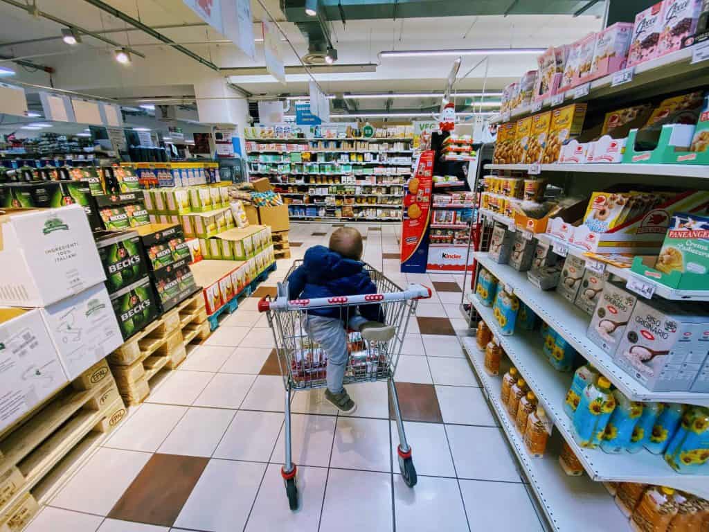 Small child sits in grocery cart in an aisle at an Italian grocery store.  You can see food on the shelves on both sides and in front of him - beer, rice, sunflower oil, and Kinder Egg products.  He is wearing grey sweatpants and a blue coat and is looking away from the camera.  