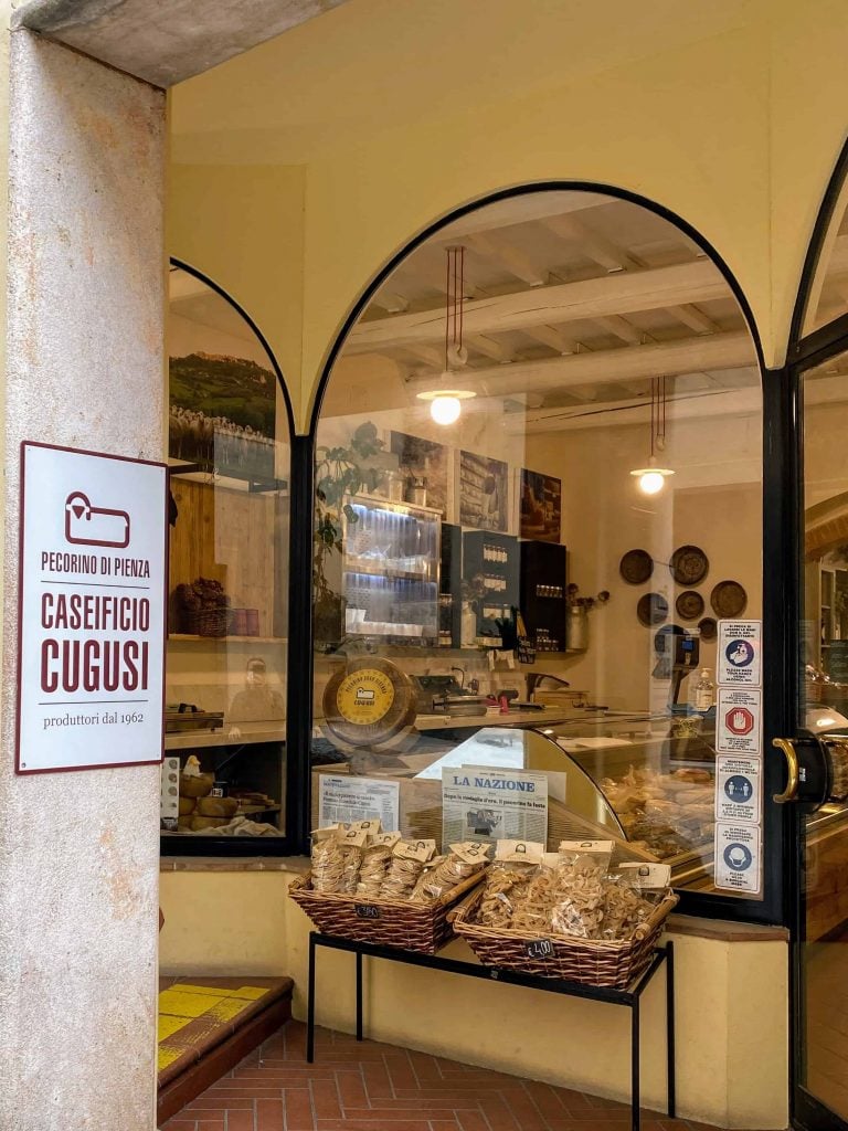 Storefront of Cugusi pecorino cheese in Montepulciano Italy.  Dried pasta is on display outside and you can see the rounds of cheese in the refrigerated display inside the shop.