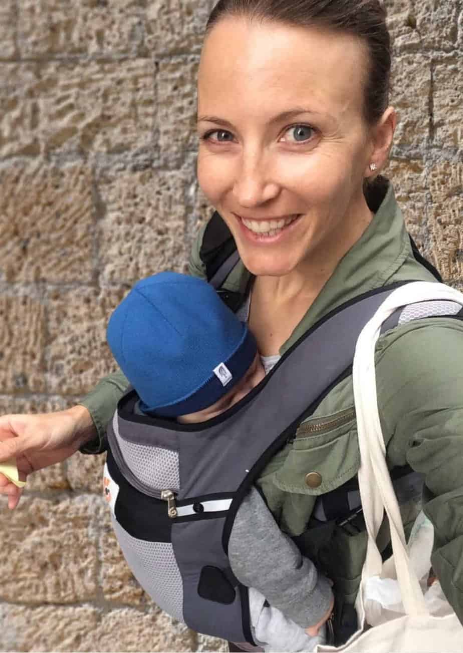 Woman carrying baby in a front baby carrier.  Baby is wearing a blue hat and is asleep.  Woman is smiling and wearing olive green jacket.