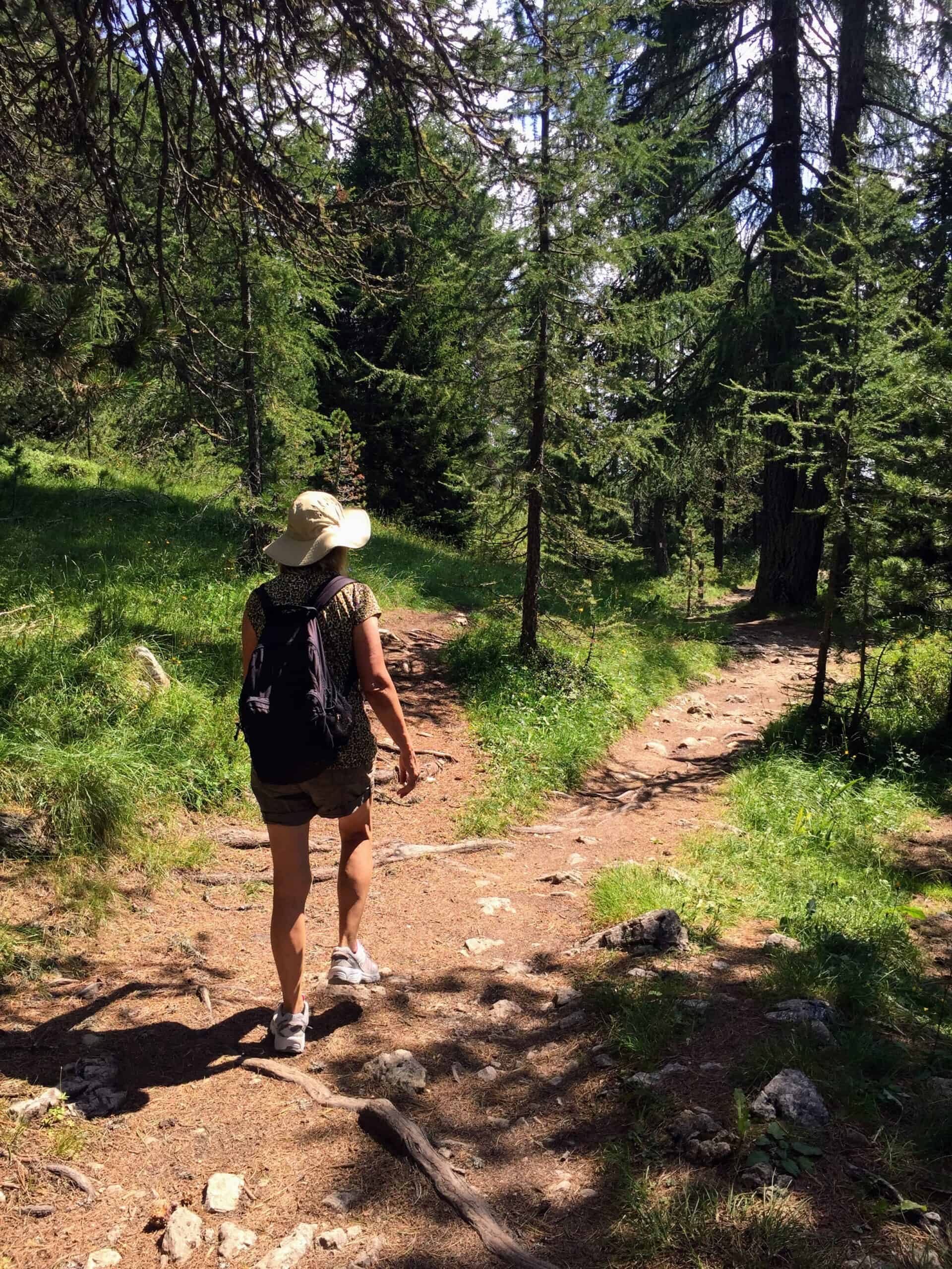 Woman hiking down the Santa Croce trail to San Cassiano in the Dolomites. You can see dirt trails and larch trees and grass.