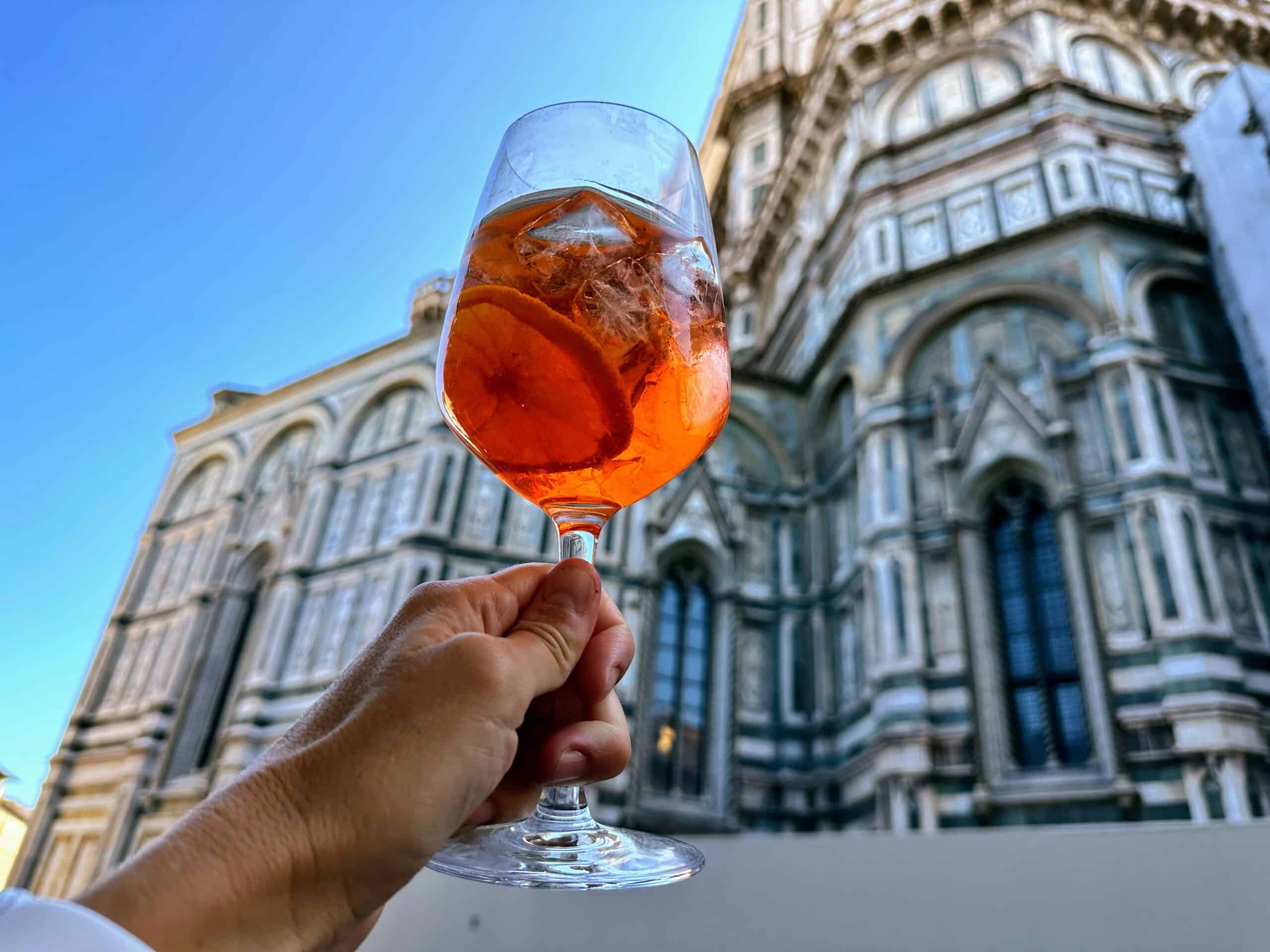 Hand holding up an Aperol Spritz in front of the Duomo in Florence, Italy.