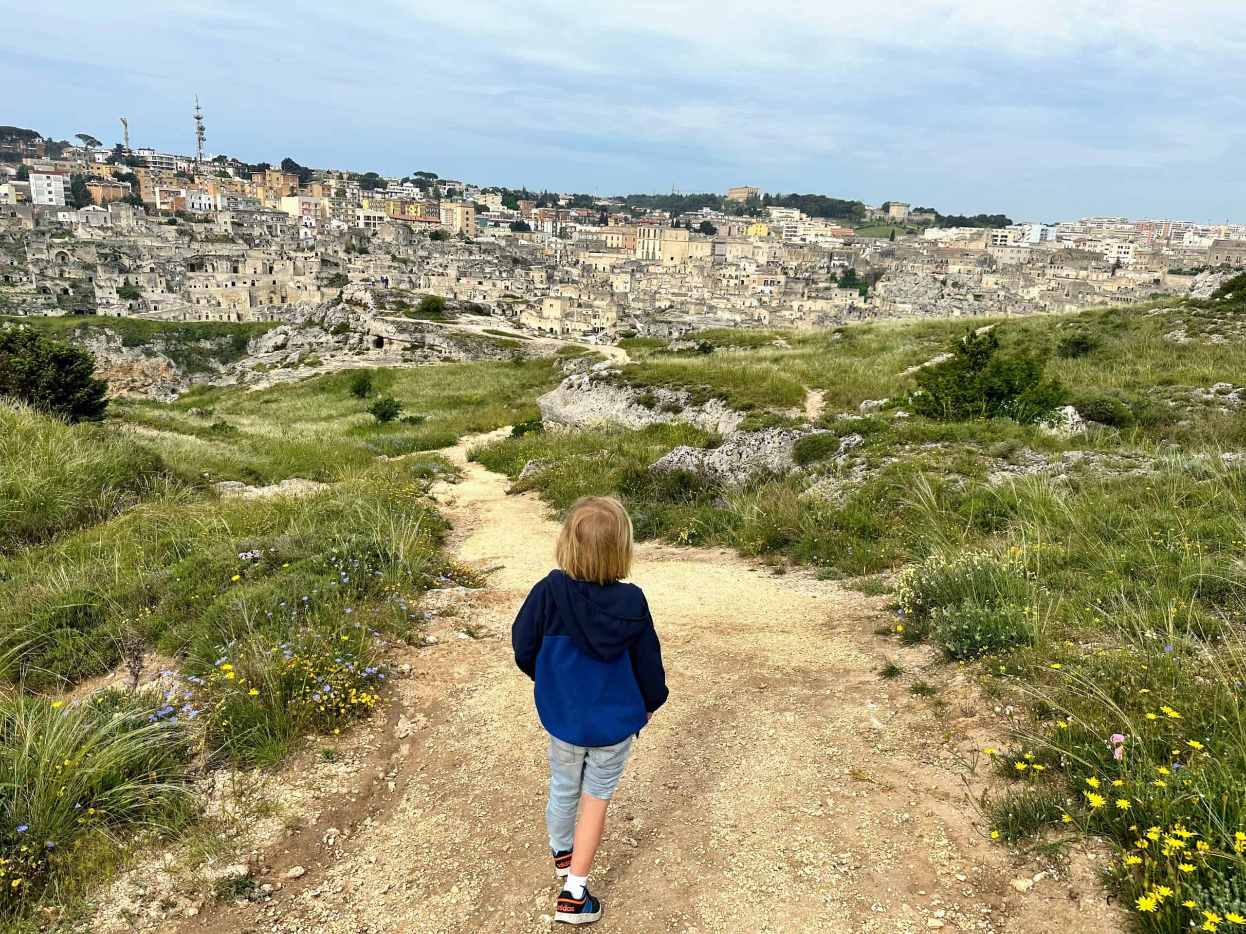 Boy hiking on dirt trail toward Matera. Grass and wildflowers on both sides of trail.