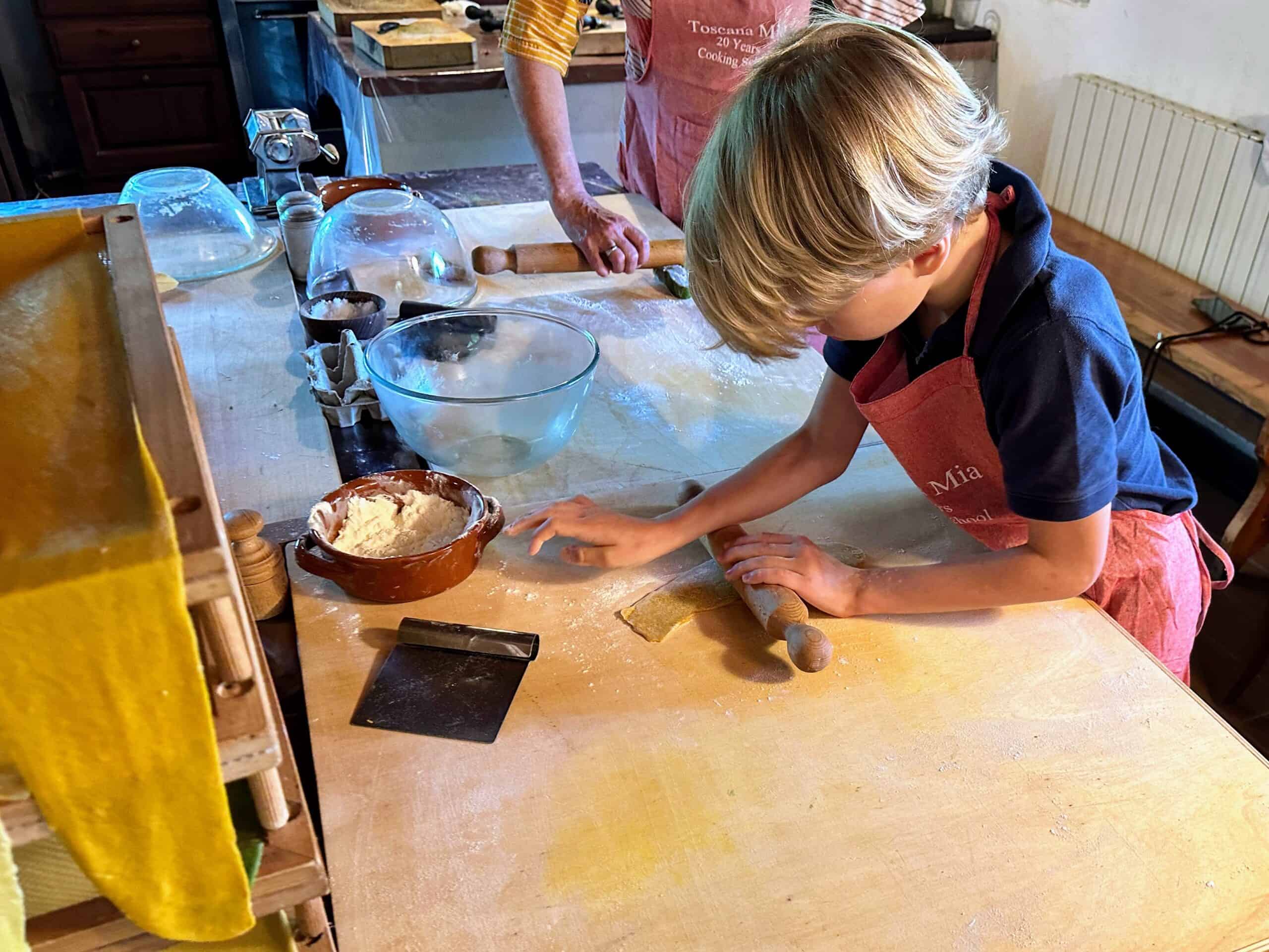 Boy leaning over wooden table and rolling fresh pasta. Some of the noodles are hanging on a rack on left.