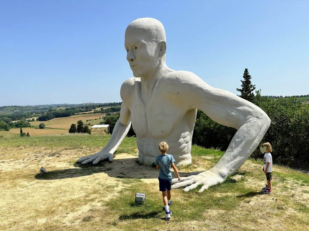 Two boys inpect the huge sculpture of a giant coming out of the ground. It's surrounded by grassy area and there are bushes and Tuscan countryside behind it.
