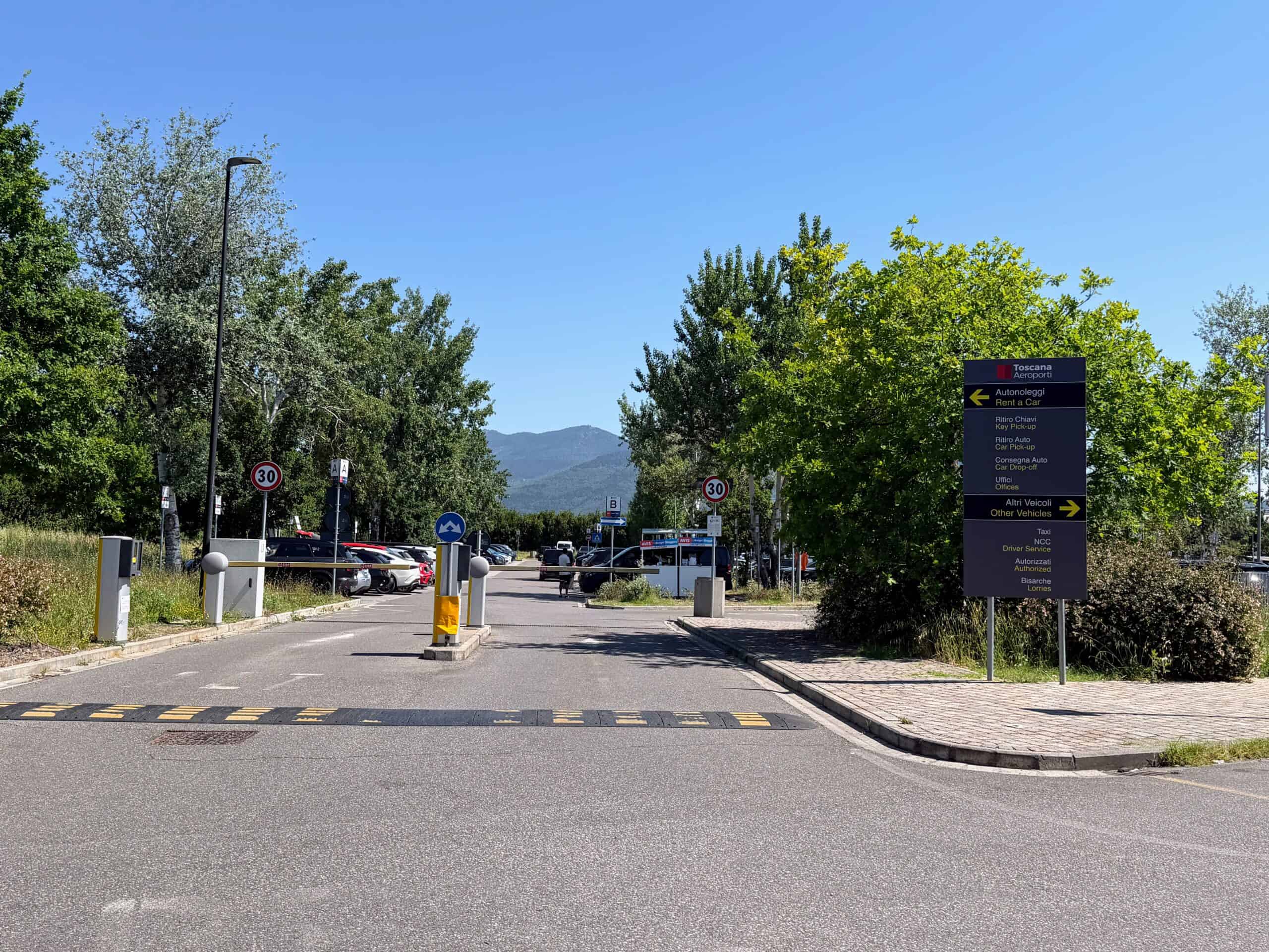 Entrance to parking lot with speed bump and barriers that can be raised. Grey sign on right. Parked cars in lot behind the barriers. Trees and bushes around the lot.