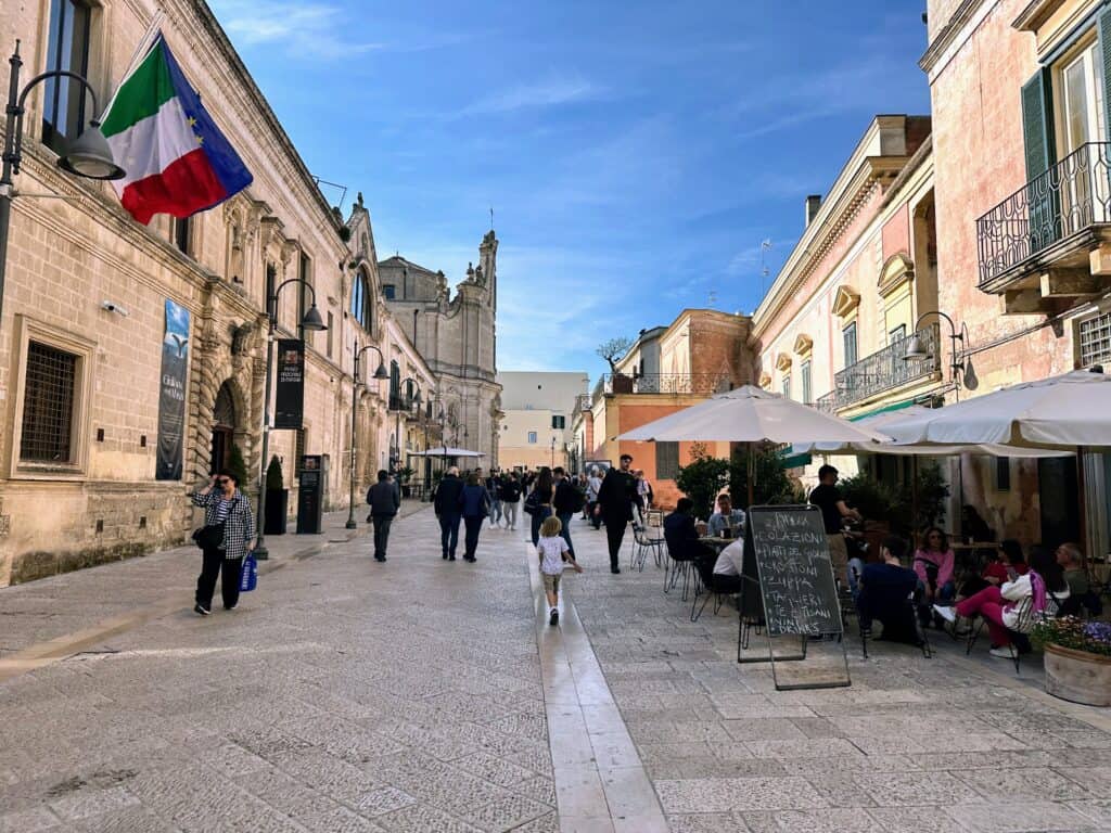 Boy walking down pedestrian street in Matera, Italy. People sitting at outdoor cafe on right. Streets are full of people walking.