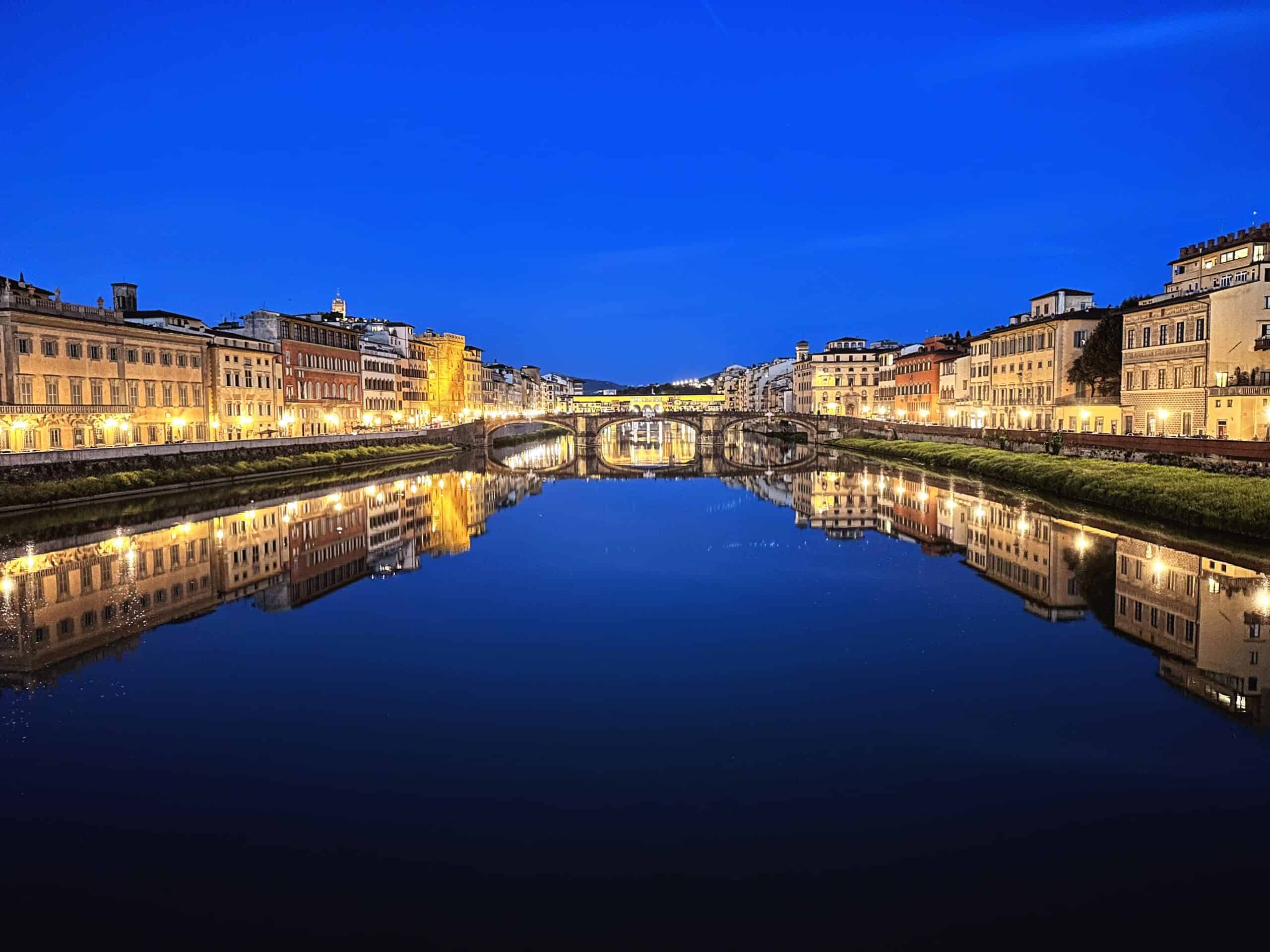 Nighttime in Florence, Italy. The Ponte Vecchio bridge is lit up and reflects on the Arno River. Lit up buildings line both sides of the river.