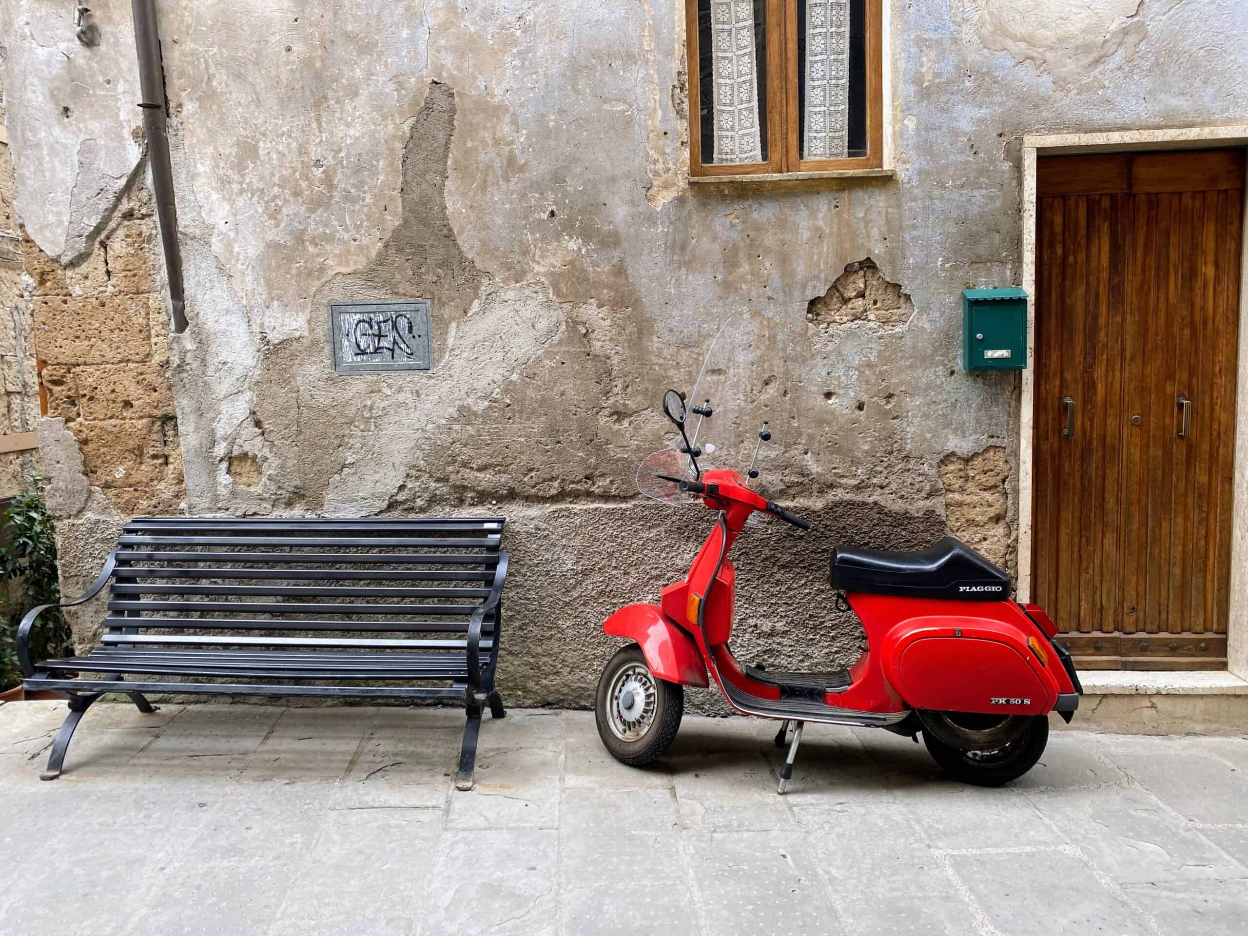 Red vespa parked against a wall in the village of Pitigliano, Tuscany. Metal bench to left and wooden door to right.