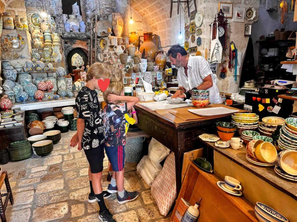 Two boys wait at the counter of a ceramics shop and the artisan wraps up their purchase of colorful bowls. The walls and shelves are decorated with ceramics.