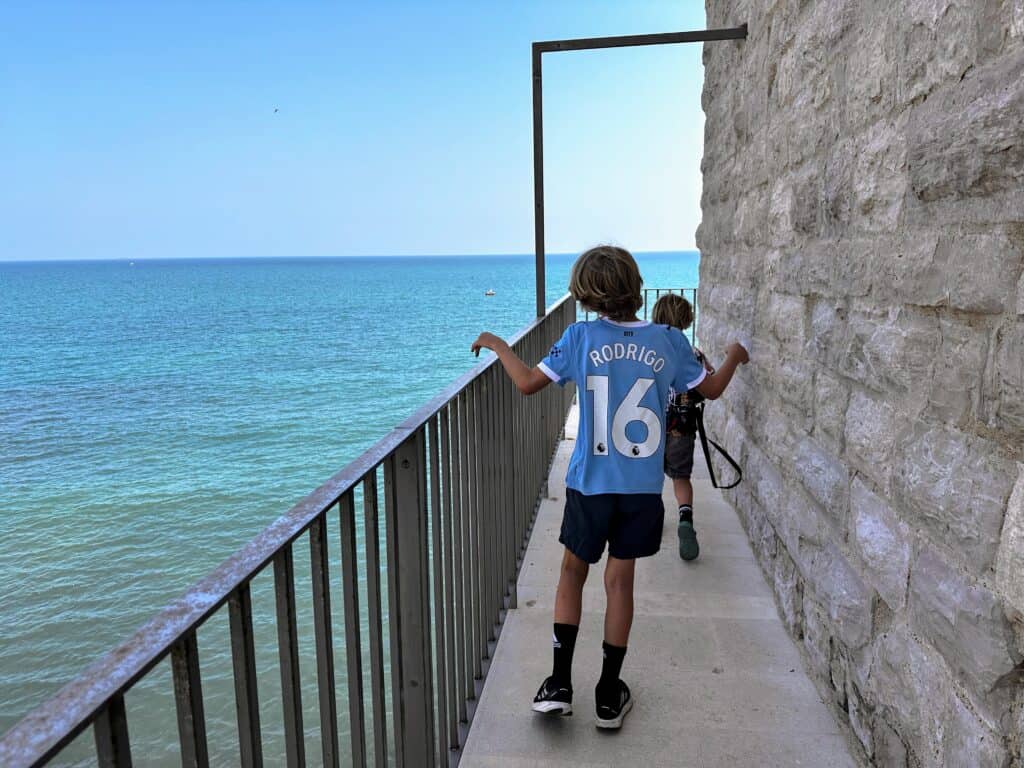 Boys walk on castle walls on the sea in Trani, Italy. Metal railing between them and the water below.