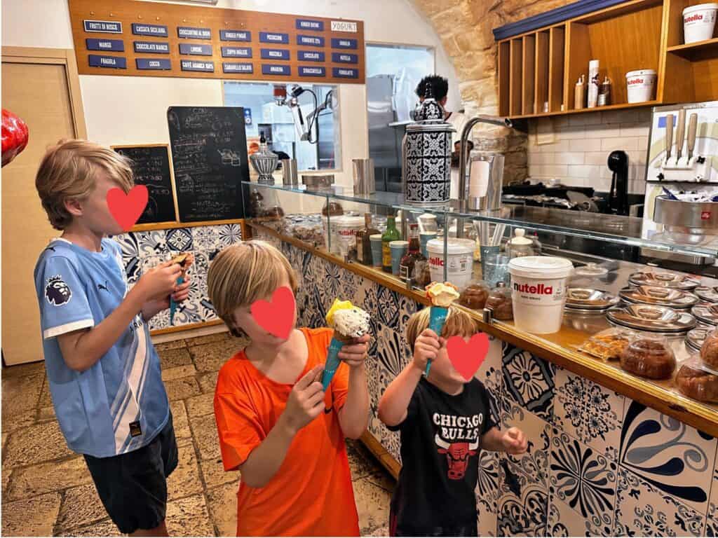 Three boys enjoy their gelato inside a gelateria in Trani, Italy. Sign with flavors on back wall and gelato in metal containers. Blue and white tiles decorate the lower counters.