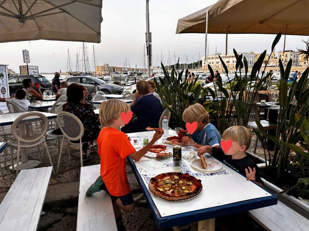 Three boys sit at a table overlooking the Trani port. They are eating pizza. The table is covered by umbrellas and there are other diners near them.