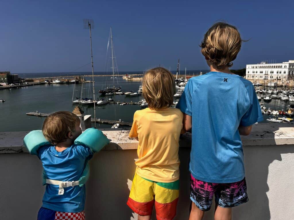 Three boys in swim gear look over a wall toward the port of Trani in Italy.