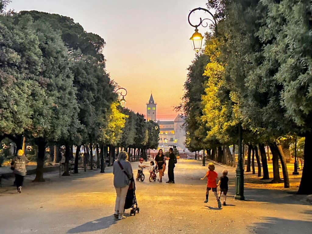 People walk down wide pedestrian lane in the Villa Comunale in Trani, Italy. It's sunset and the buildings in the distance are lit up in golden colors.