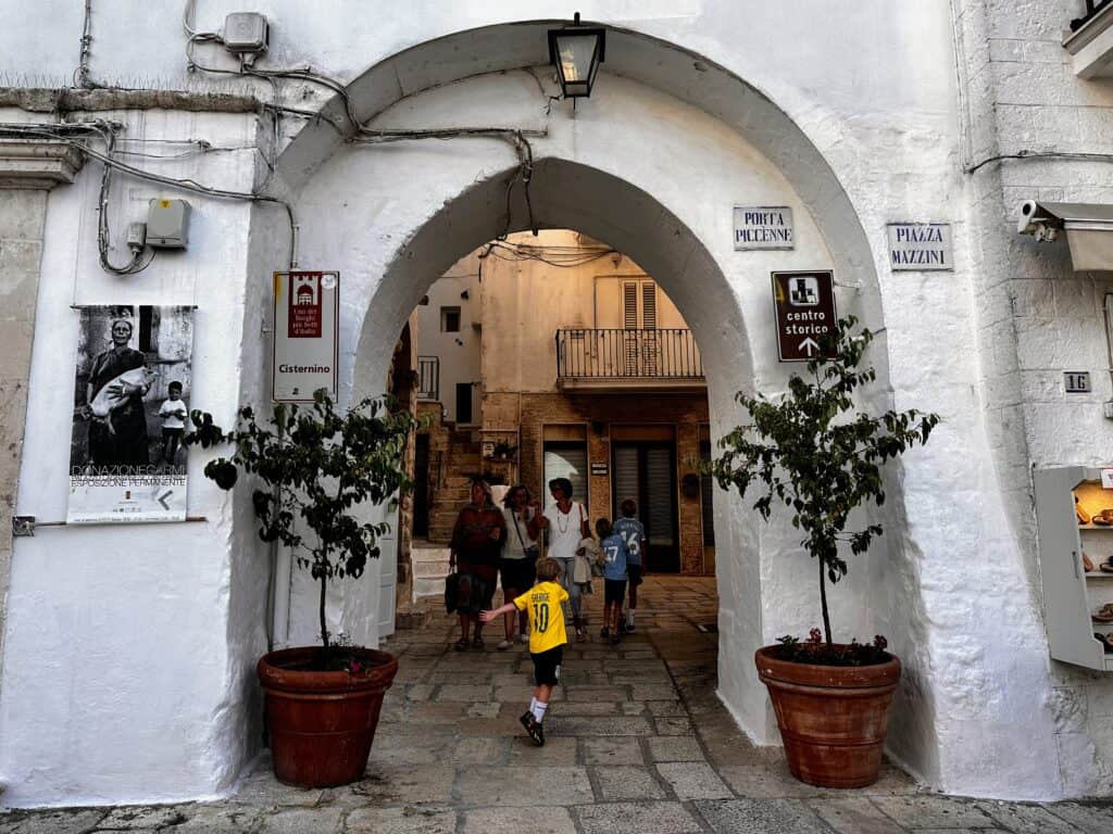 Boys walk under an archway to enter the historic center of Cisternino in Puglia, Italy. Trees in terracotta pots on either side. Sign for Piazza Mazzini and Porta Piccènne on the right of the arch. Three women walk out of the arch toward the camera.