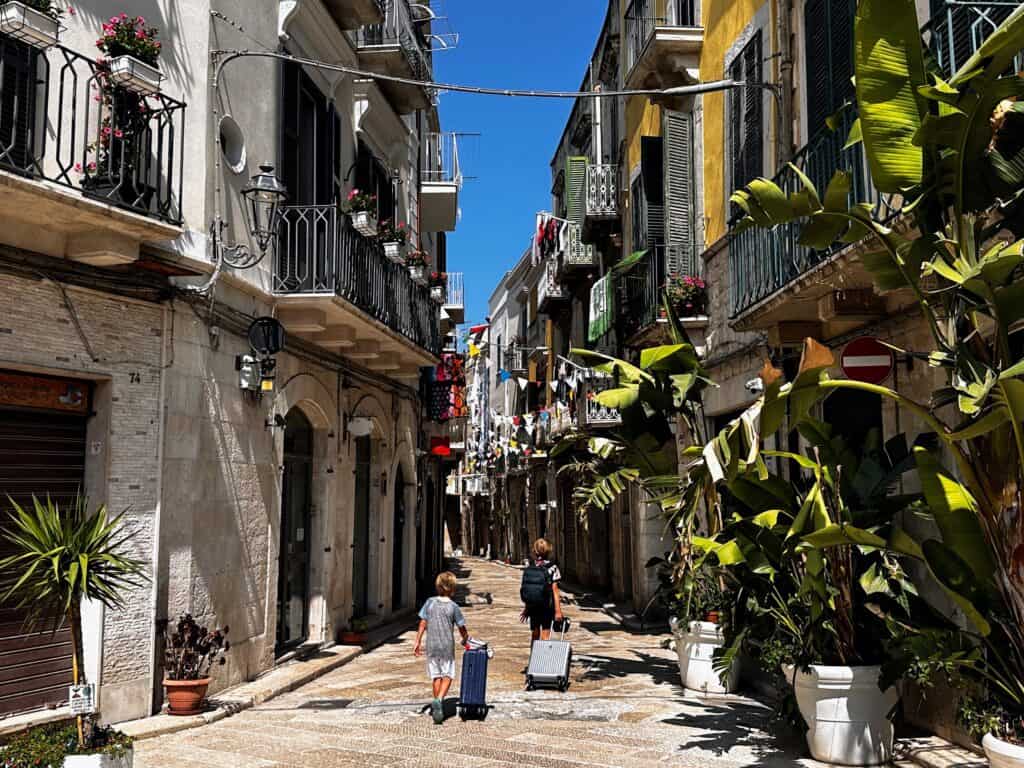 Two boys pull carry on suitcases down a stone street in Trani, Italy. Plants on the right, balconies on the multi-story buildings.