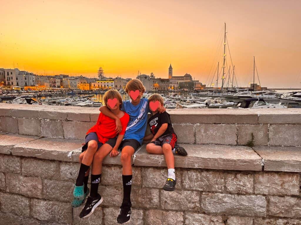 Three boys sit on a stone wall with the port of Trani and the sunset behind them.