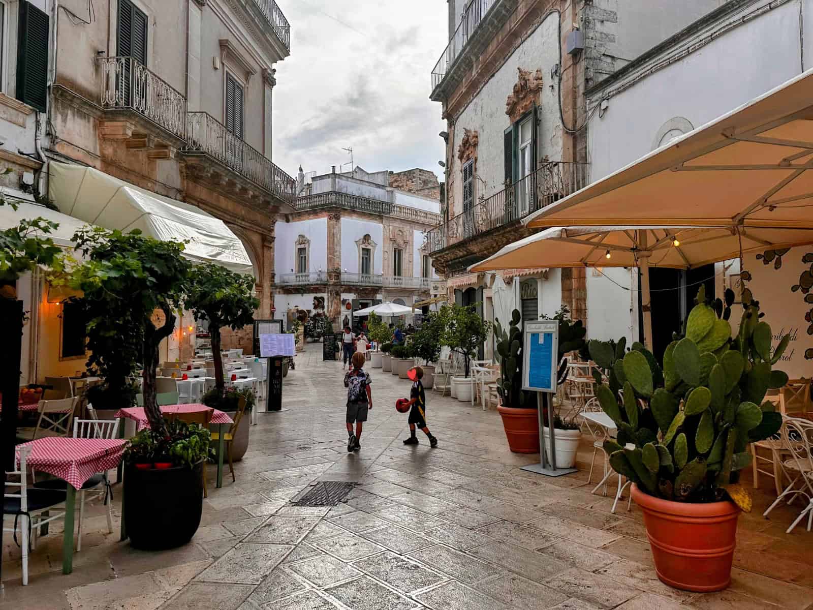Two boys walk down a stone street in a pedestrian area of Martina Franca, Puglia. Restaurants with outdoor tables lines both sides of the street. Old buildings on either side with balconies with iron railings.