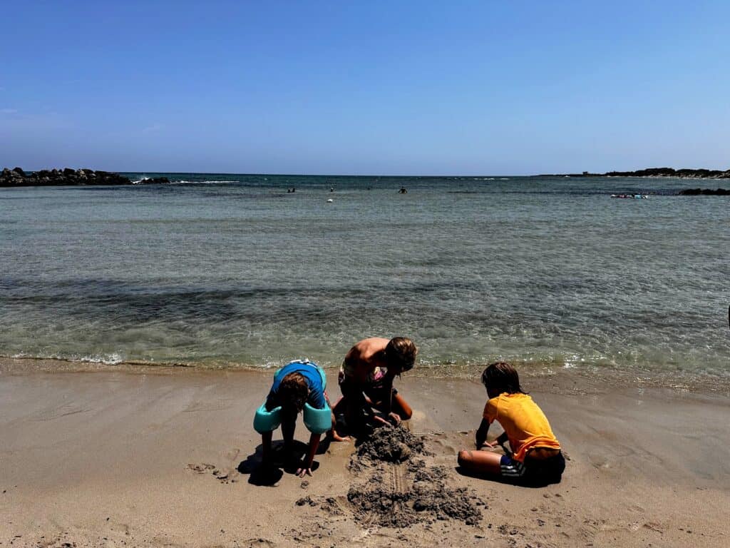 Three boys play in the sand at the edge of clear, calm water at the Torre Guaceto beach in Puglia, Italy.