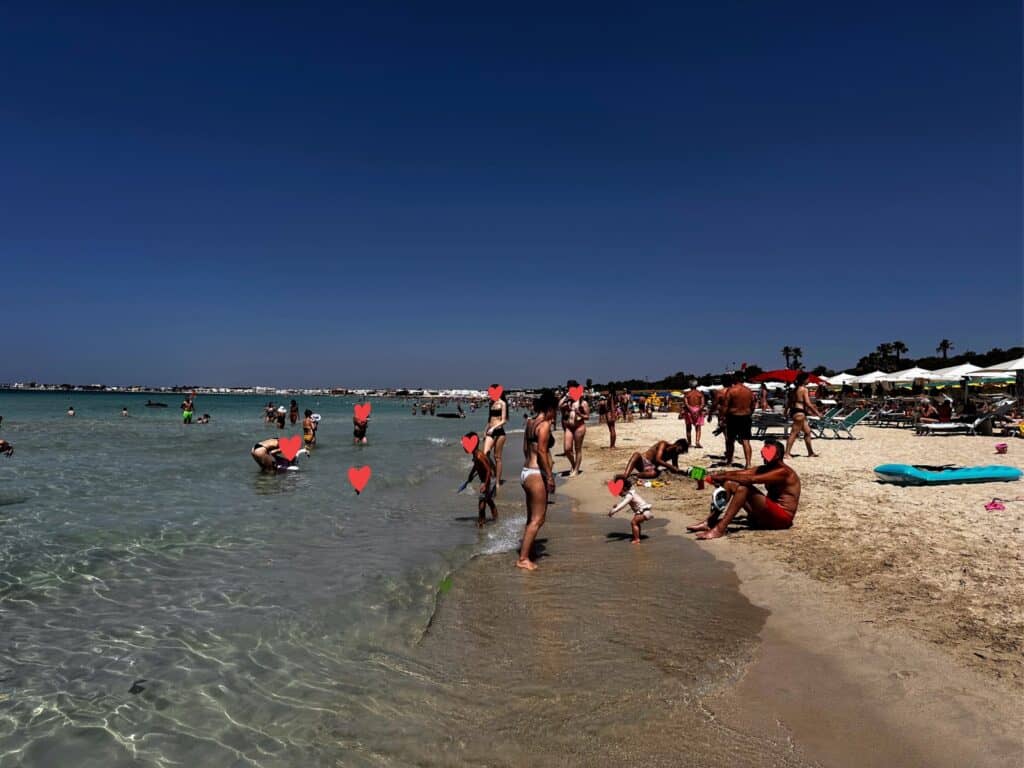 People play in the sand and the shallow water at Lido Stella Maris in Puglia. White beach umbrellas on edge of sand on right.