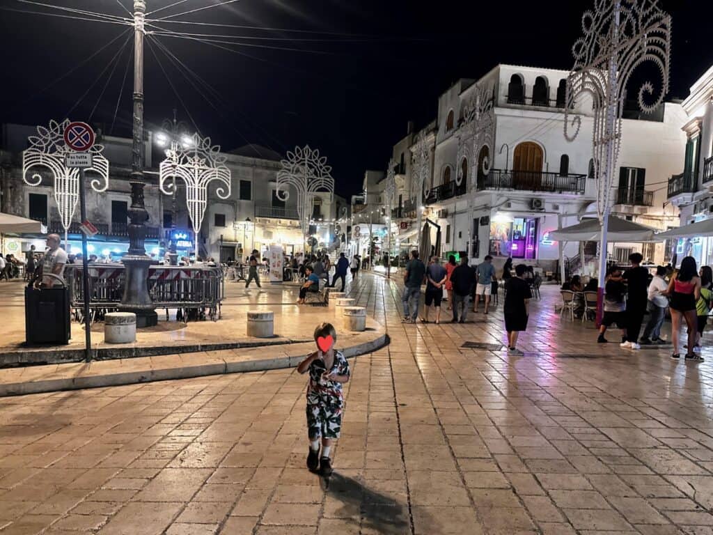 Boy in floral shirt and shorts set is eating ice cream and walking in Piazza Plebiscito in Ceglie Messapica. The piazza and streets around it are decorated with large white luminarie. There are people walking around and sitting at cafes on the edge of the square.