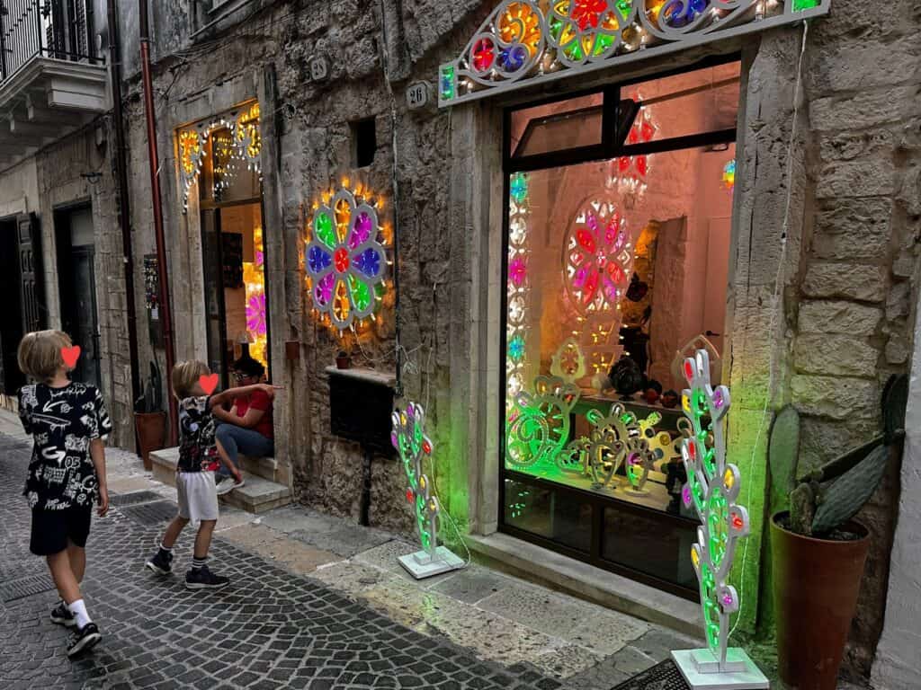 Two boys walk on a cobblestone pedestrian street and one points at a shop window full of colorful luminarie. They're in Ceglie Messapica in Puglia, Italy.