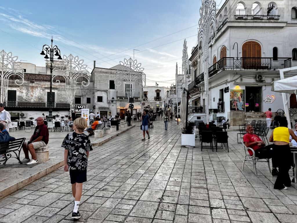 Boy walks on a pedestrian street in the center of Ceglie Messapica. People sit at sidewalk cafes. Luminarie decorate the edges of the piazza.