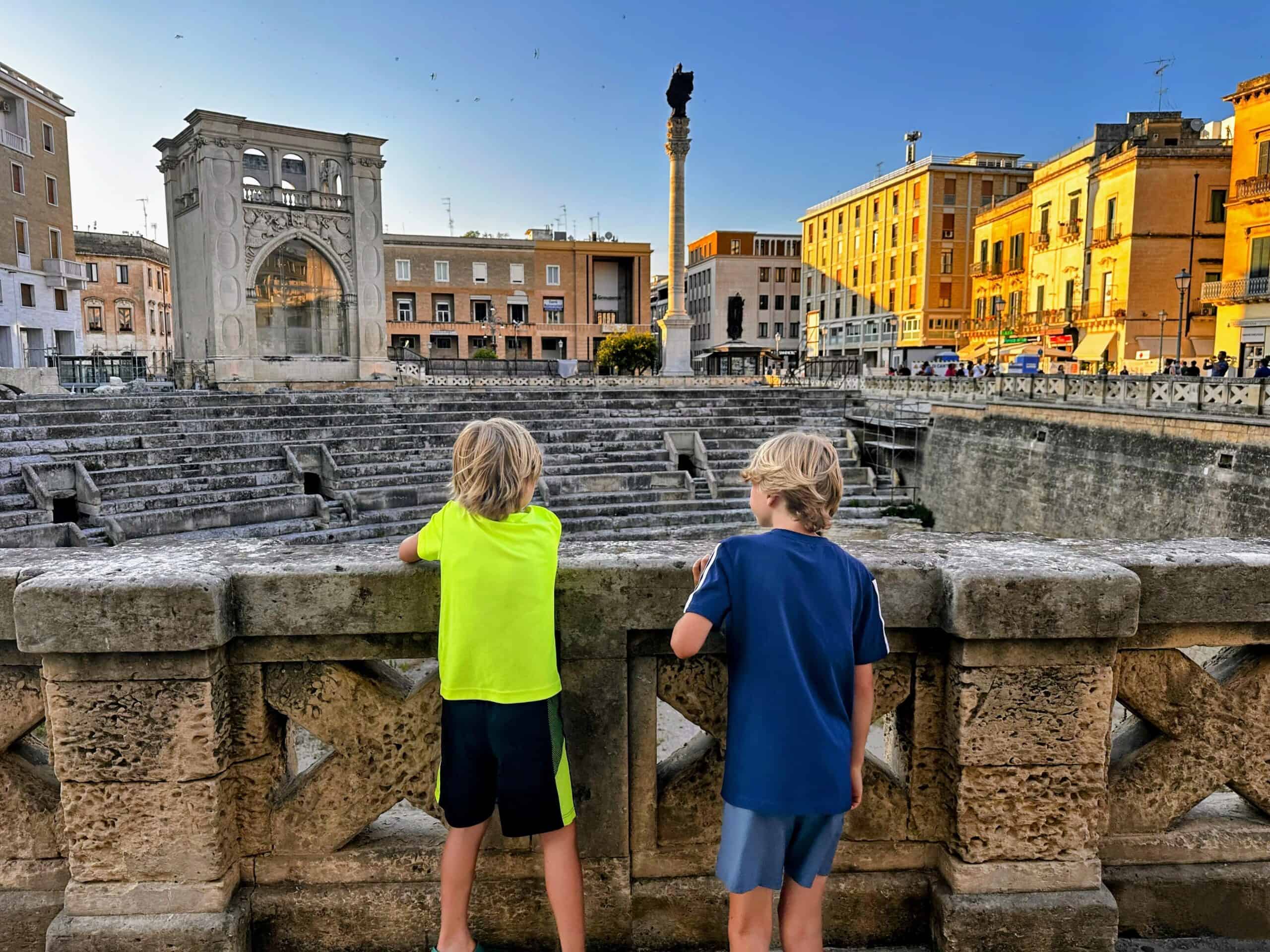 Two boys look over stone railing into the Roman amphitheater below them. They're in Lecce, Puglia. Stone buildings in the background and birds fly in the sky.