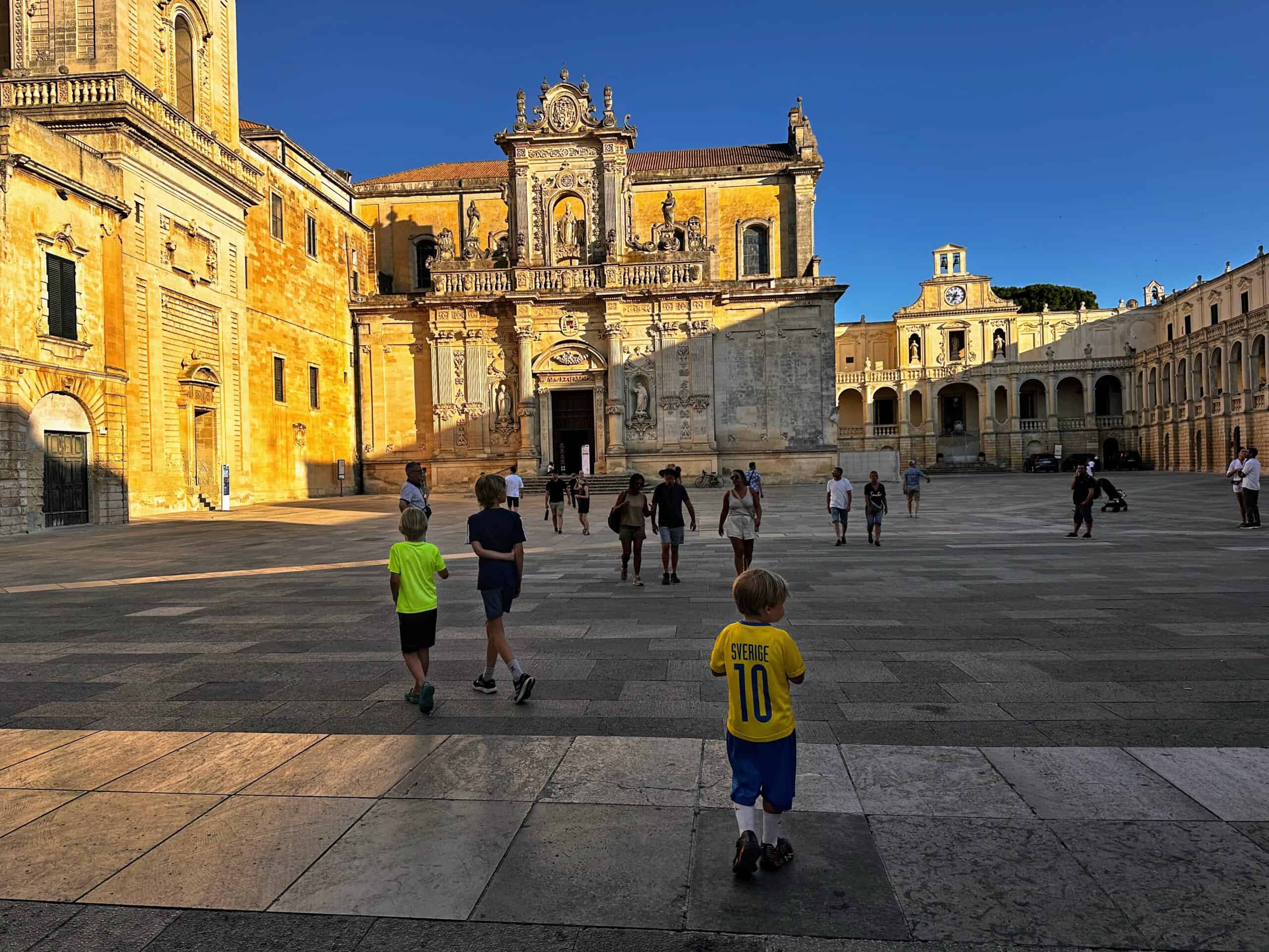 Three boys walk into a large square in Lecce, Puglia. Church ahead and others are walking in the square but it's not crowded. The sun is setting so part of the square is shaded.