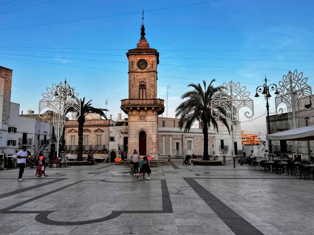 A few kids and adults play in front of a clock tower on a stone piazza in Ceglie Messapica, Puglia. Luminarie and palm trees on either side of the clock toer.
