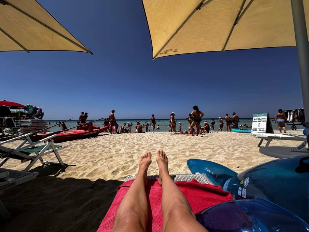 Woman's legs on lounge chair at beach overlooking people at the water's edge. Taken at Torre Lapillo beach in Puglia, Italy.