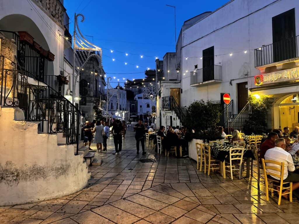 Lights hang above the small pedestrian street at night in Ceglie Messapica in Puglia. People dine outdoors and chat on the street.