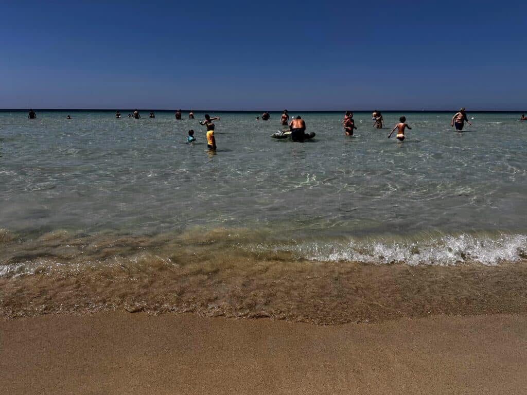 People standing in shallow water at Torre Lapillo beach in Puglia. Small wave crashes in the sand.