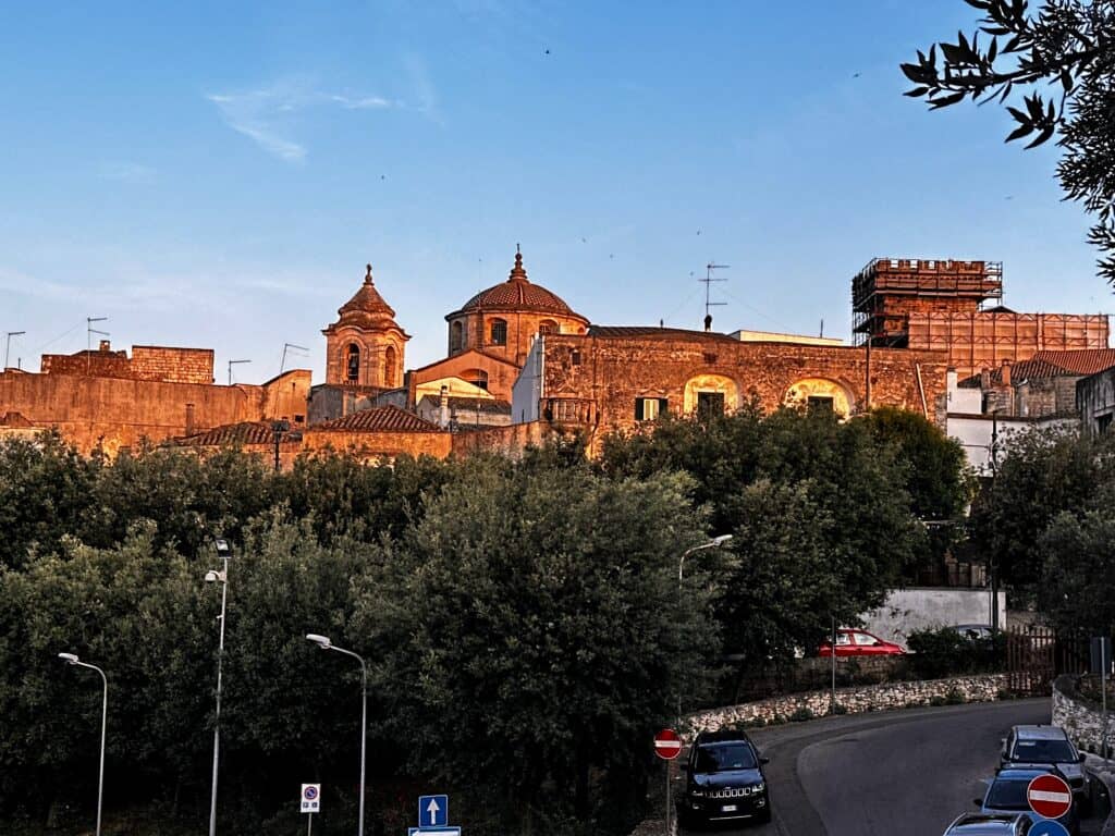 Rooftops of Ceglie Messapica in Puglia, Italy look red as the sun sets. Large trees in front of them and some cars parked on a street.