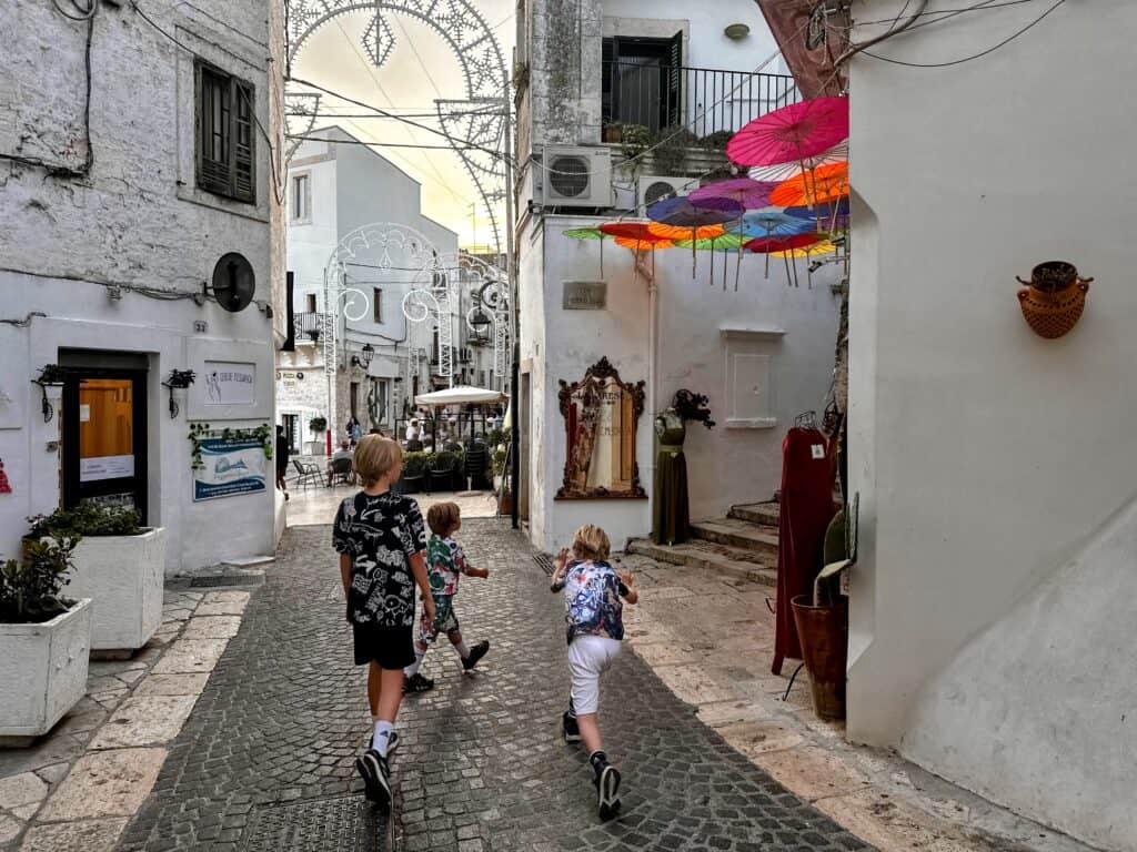 Three boys walk on a cobblestone street in a small town in Puglia (Ceglie Messapica). White walls, colorful umbrellas decorate between buildings on the right. In front of them the street opens onto a piazza with an outdoor cafe.