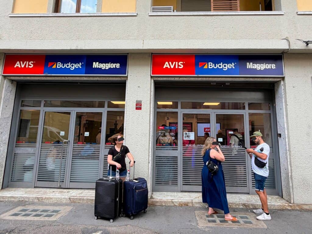 People waiting outside and inside the car rental office for Avis, Budget, and Maggiore in Florence, Italy. One woman out front holds two suitcases.