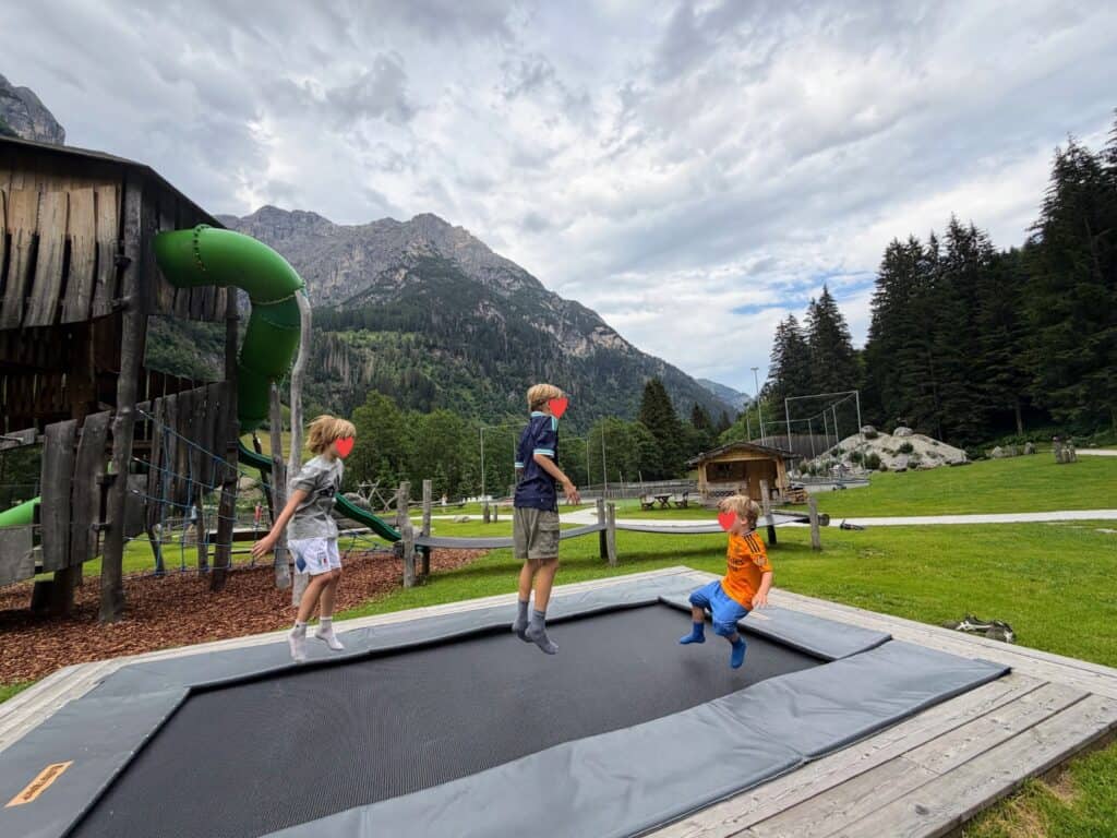 Three boys jumping on an outdoor trampoline. It's set into the ground and to the left is a wooden play structure with green tube slide and bumpy slide. Mountains, pine trees, and soccer pitch behind them.