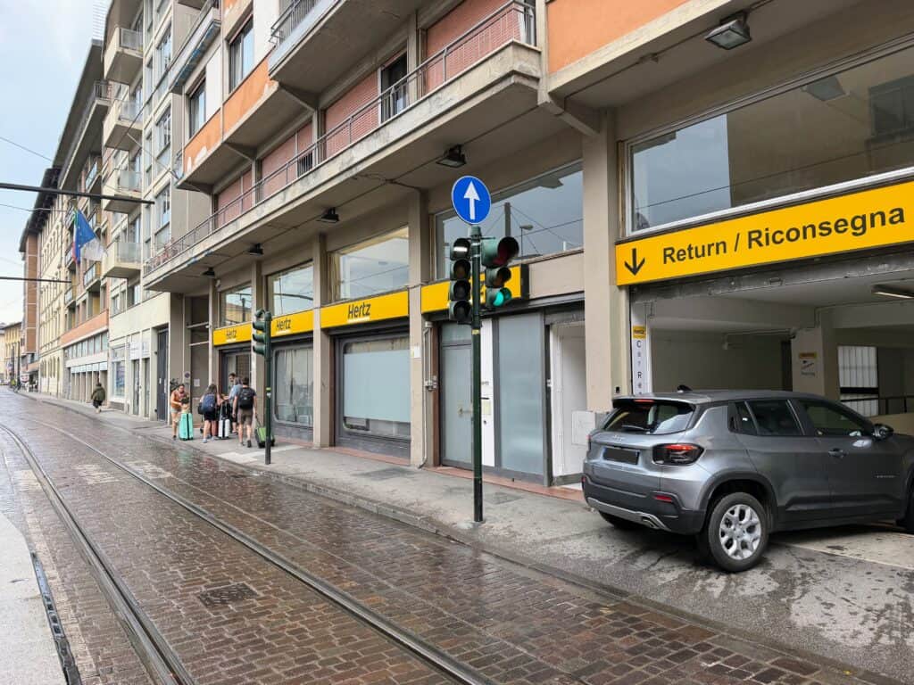 Grey car drives down into a parking garage entrance off of a cobblestone street in Florence, Italy. The street has tram lines. The sign on the building says Hertz and Return/Riconsegna. There is a traffic light and a few people waiting with suitcases under the sign for Hertz.