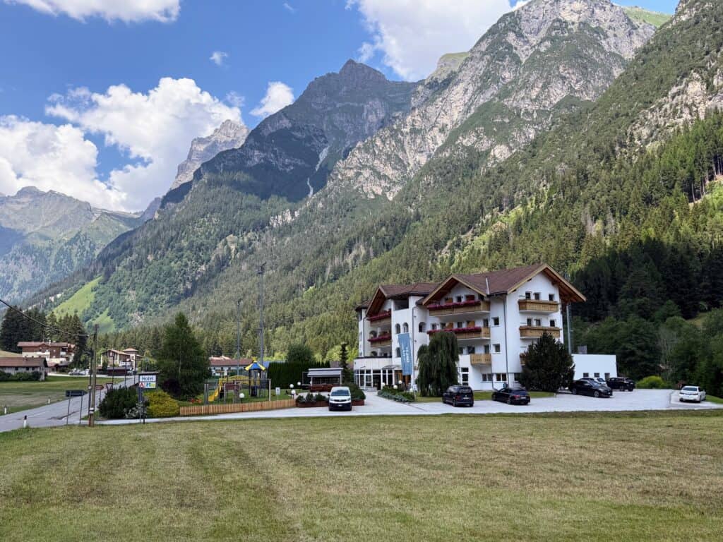 White, four-story hotel set at the base of the Dolomite mountains. Road to the left. A few cars parked in the hotel driveway. Sunny day with puffy white clouds.