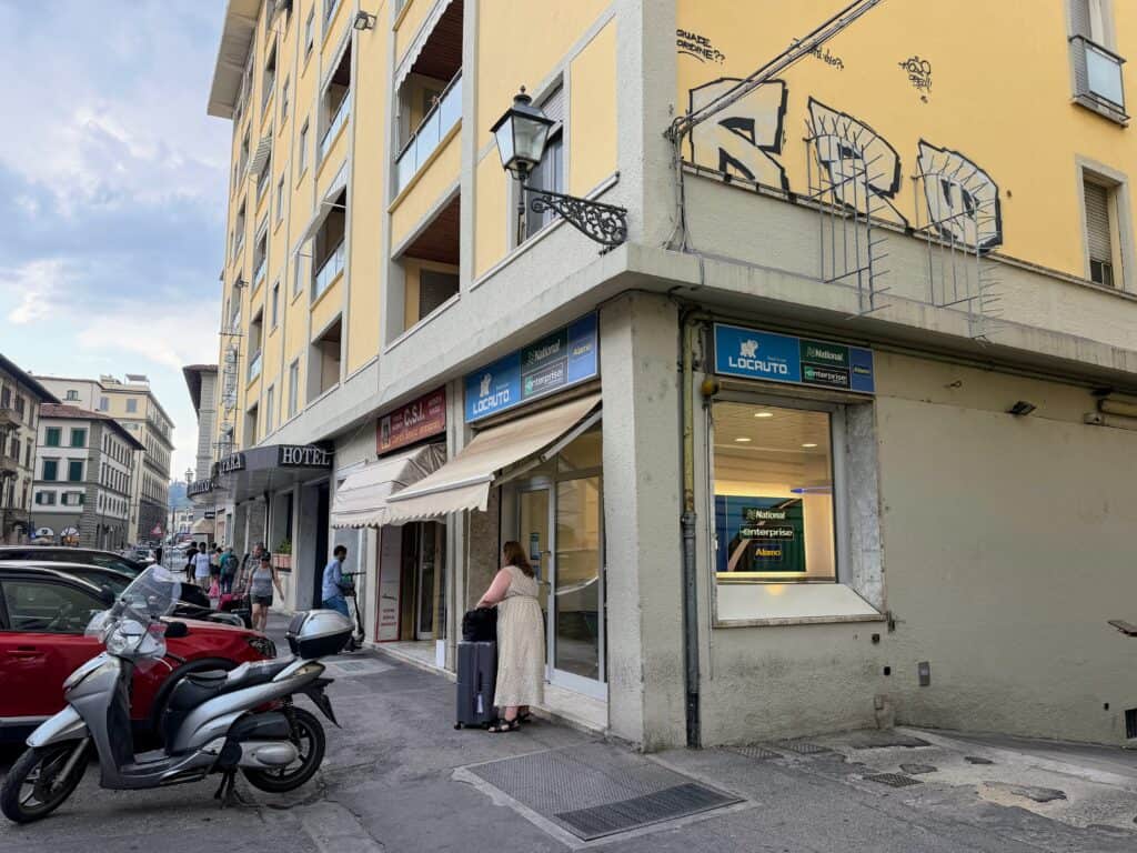 Corner street in Florence, Italy. Yellow multi-story building with graffiti on the side. On left there are motorcycles and cars parked. The lower floor of the building is the car rental office for Locauto, National, Enterprise, and Alamo car rental companies. This is in Florence, Italy.