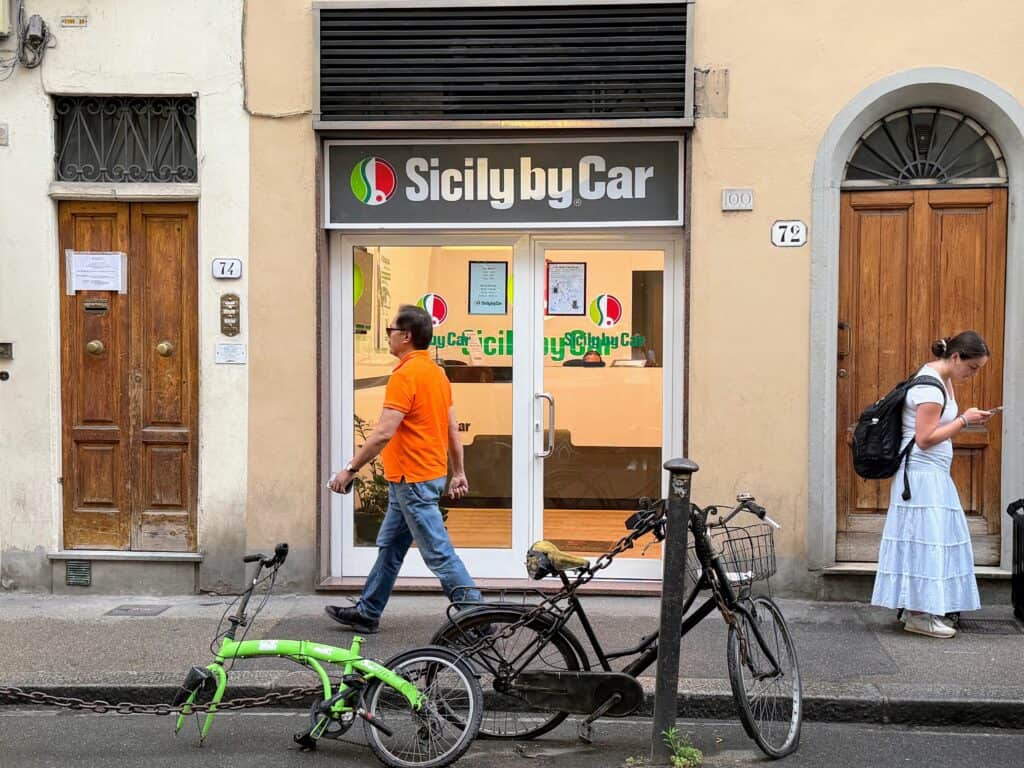 Front office of Sicily by Car car rental agency in Florence, Italy. Glass entrance and man working at desk inside. Out front, a man in an orange shirt walks by the office entrance and a woman in a white dress looks down at her phone. There are two bicycles parked on the street out front.