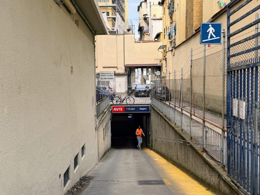 Man in orange shirt walks up driveway entrance to the car rental garage for Avis, Budget, and Maggiore. It's on a lower level and above it are cars parked and a bicycle.