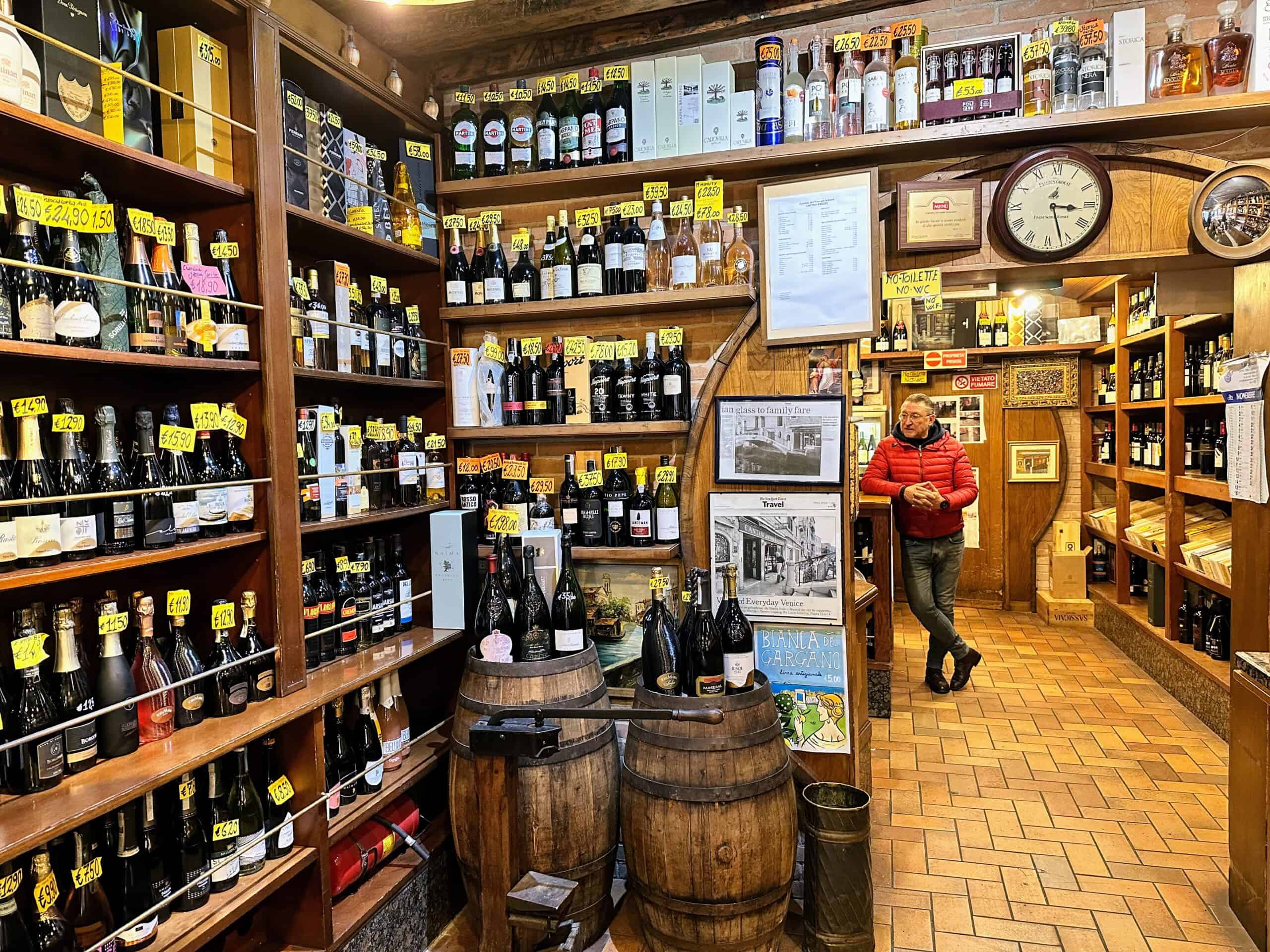 Man stands at counter in back of small bar in Venice, Italy. Shelves lined with wines and liquors.