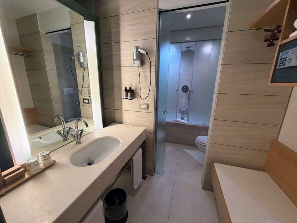 Light colored hotel bathroom at Precise House Mantegna Rome. Sink area on left, dressing area with hangers and safe on right. In back is toilet and shower/bath.