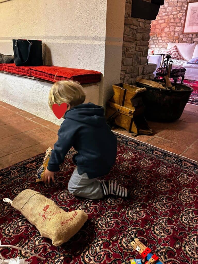Boy opens up his Befana stocking on the floor in a house in Italy. Living room furniture in room to his right.