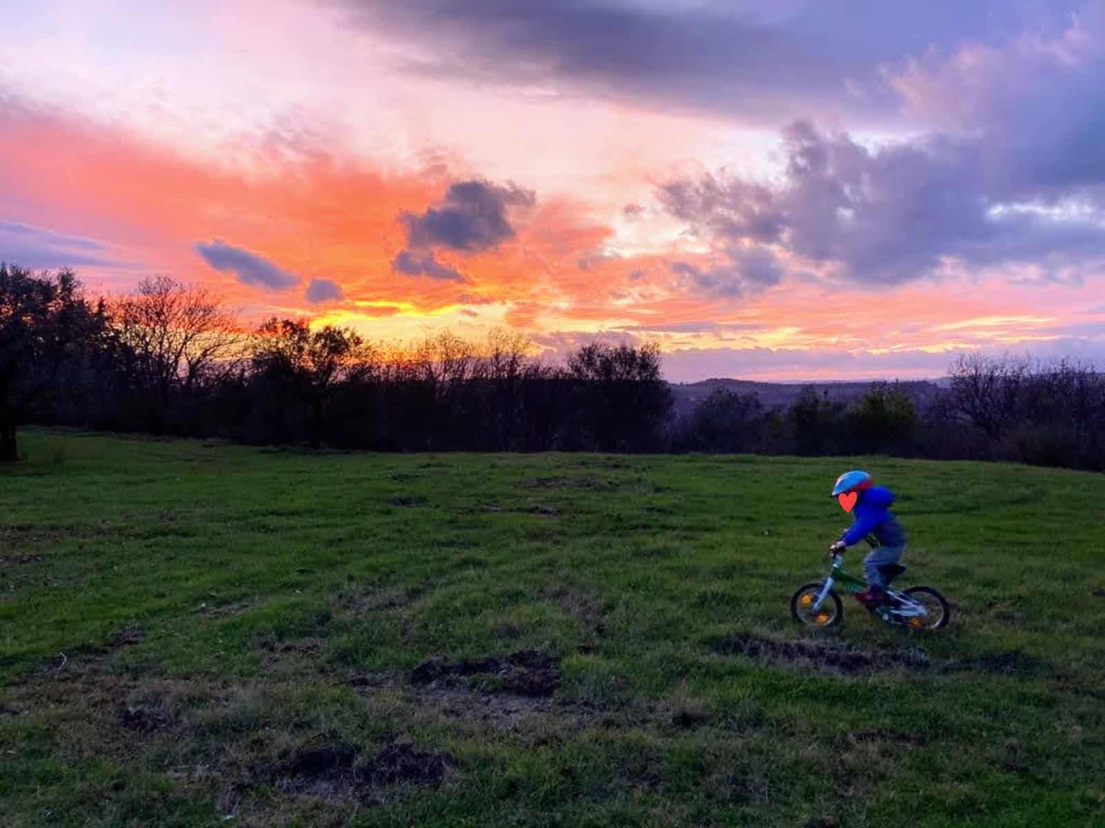 Boy riding bike in grass in Tuscany. Pink and orange sunset behind him.