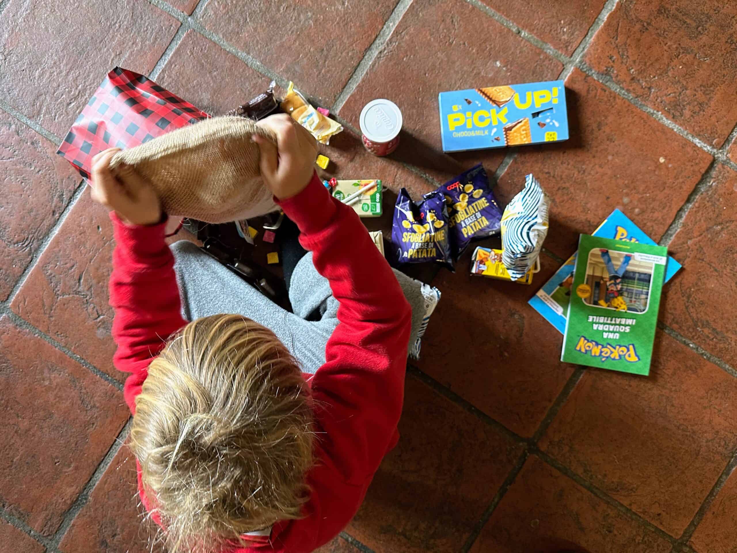 Boy opens up Befana stockings on floor in home in Italy. Snacks and books come out.